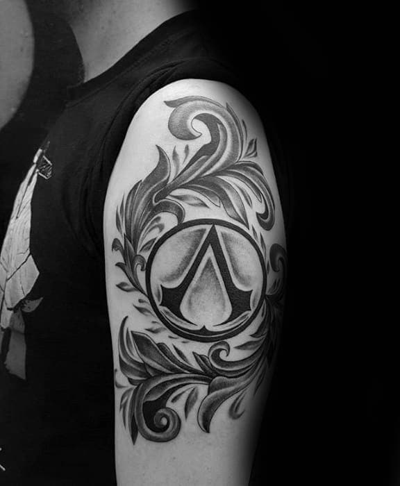 Symbol Assassin’s Creed z ornamentem na ramieniu Czarno-szary tatuaż z symbolem Assassin’s Creed i ornamentalnymi liniami na ramieniu